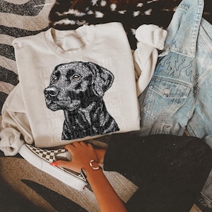 May include: A cream-colored sweatshirt featuring a detailed black and silver glitter Labrador Retriever graphic. The dog's portrait is centered on the front, with text partially visible. A denim jacket and checkered sneakers are also in the image.