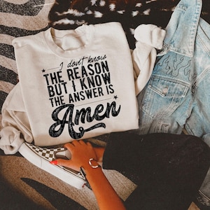 May include: A cream-colored sweatshirt with the text "I don't know the reason but I know the answer is Amen" in black. A denim jacket with flame designs and a pair of checkered sneakers are also visible.