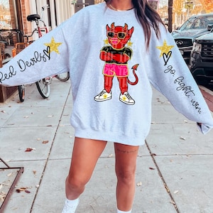 May include: A light gray sweatshirt with the words "Red Devils" on the left sleeve and "go. fight. win." on the right sleeve. The front features a cartoon devil wearing sunglasses and shorts. Gold stars and hearts accent the sleeves.