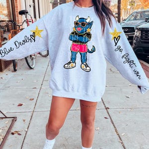 May include: A light gray sweatshirt featuring a cartoon devil character wearing sunglasses and pink shorts. The sleeves have the phrases "Blue Devils" and "go. fight. win." with gold star and heart accents. The sweatshirt is a casual style.
