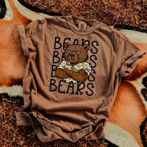 May include: A brown t-shirt with the word "BEARS" stacked vertically and a cartoon bear graphic. The bear is wearing a collared shirt and has its arms crossed. The shirt has rolled-up sleeves.