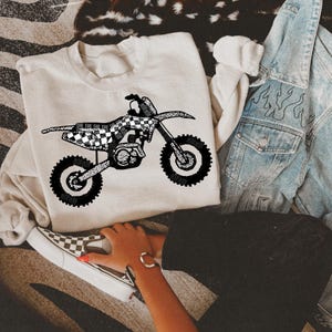 May include: A white sweatshirt with a black and white checkered dirt bike graphic. The bike is made up of the words "The" and "Ride".