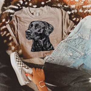 May include: A tan t-shirt featuring a black Labrador Retriever design. The dog's image is made of sparkling material. The shirt is paired with a light blue denim jacket with flame designs and checkered sneakers.