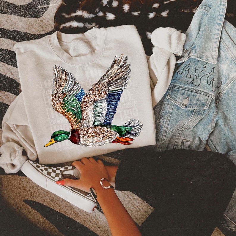 May include: A light gray sweatshirt with a colorful sequined mallard duck design. The duck is flying with its wings spread out. The sweatshirt is being worn by a person with a black shirt and jeans.