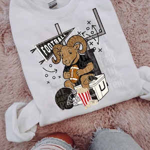 Pode incluir: Camisola branca com um gráfico de um carneiro segurando uma bola de futebol, um capacete e pipocas. A palavra "FOOTBALL" está em uma faixa. O texto "The Classy Trio" também é visível.
