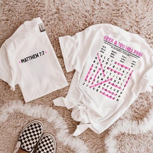May include: Two white t-shirts. The left shirt features the black text "MATTHEW 7:7" flanked by two pink stars. The right shirt displays the pink text "SEEK & YOU WILL FIND" above a word search grid with pink highlighted words and a large pink X.