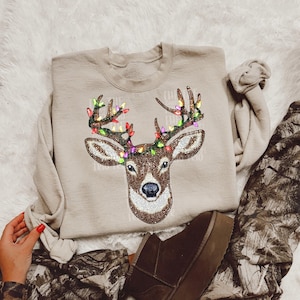 May include: Beige crewneck sweatshirt featuring a deer head design. The deer's antlers are adorned with colorful Christmas lights. The deer's head is detailed with a pattern of small, sparkling embellishments. The sweatshirt is a neutral color.