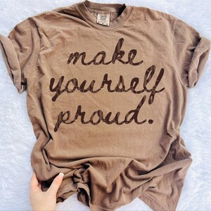 Puede incluir: Una camiseta marr&oacute;n con la frase "make yourself proud" en marr&oacute;n oscuro, con fuente cursiva. La camiseta tiene cuello redondo y mangas enrolladas. La tela parece suave y c&oacute;moda.