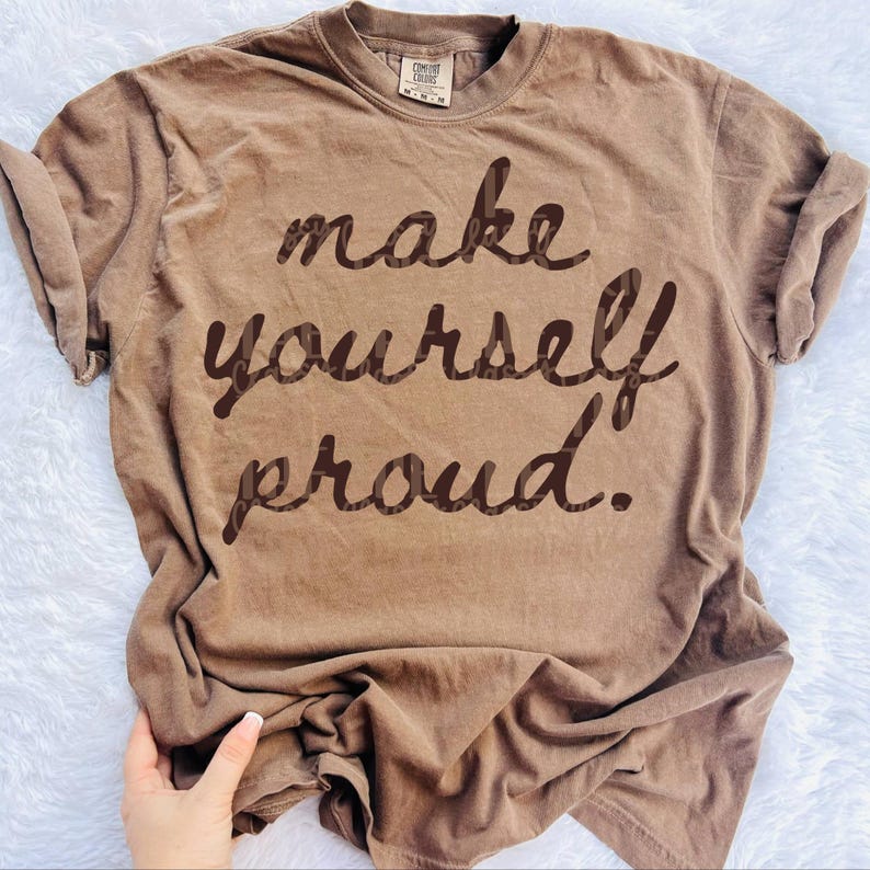 Puede incluir: Una camiseta marr&oacute;n con la frase "make yourself proud" en marr&oacute;n oscuro, con fuente cursiva. La camiseta tiene cuello redondo y mangas enrolladas. La tela parece suave y c&oacute;moda.
