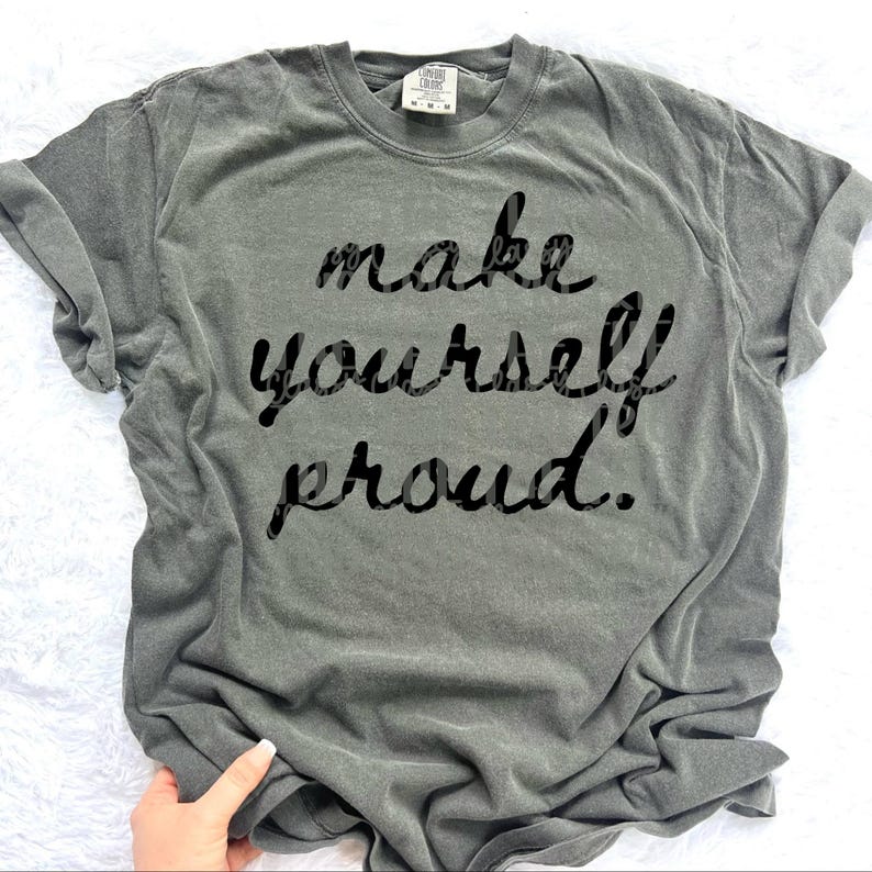 Puede incluir: Una camiseta gris con la frase "make yourself proud" en negro, con una fuente cursiva. La camiseta tiene mangas cortas y cuello redondo. El texto tiene un estampado de leopardo.