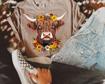 Puede incluir: Una vaca Highland marrón y blanca con una corona de flores en la cabeza. La vaca está en una camiseta marrón claro.