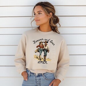 Puede incluir: Una sudadera beige con un estampado gr&aacute;fico de una pareja de vaqueros caminando. El texto en la sudadera dice "Forever and Ever Amen".