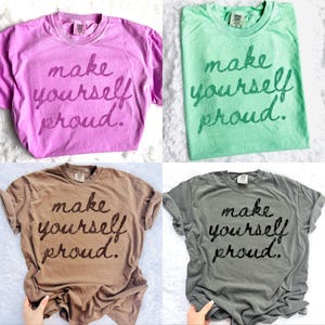 Puede incluir: Cuatro camisetas de manga corta en tonos rosa, verde, marr&oacute;n y gris. Cada camiseta muestra la frase "make yourself proud" en un estilo manuscrito. Las camisetas est&aacute;n dispuestas sobre una superficie blanca texturizada.