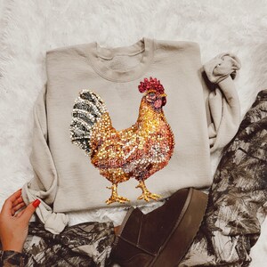 May include: A beige sweatshirt with a sequined rooster design. The rooster is brown, gold, and black with a red comb.