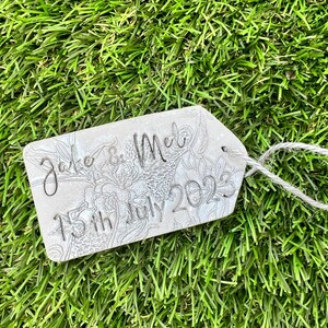 May include: A white tag with a floral design and the text "Jake & Mel 15th July 2023" on it. The tag is tied with a white string and is sitting on a green grassy surface.