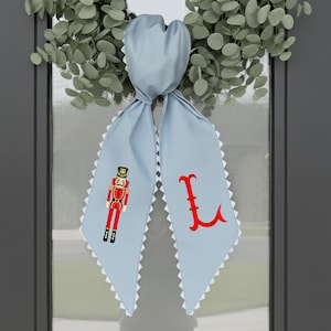 Puede incluir: Un adorno de puerta azul claro con un borde festoneado blanco. Un lado presenta un soldado cascanueces rojo y negro, el otro una letra roja "L". El adorno está atado en la parte superior y cuelga frente a una corona verde.