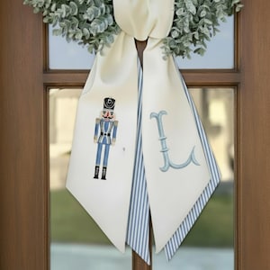 Puede incluir: Faja de puerta color crema con borde a rayas azules y blancas y una figura decorativa de cascanueces. La faja también presenta una letra "L" bordada en azul claro y está adornada con una corona verde.