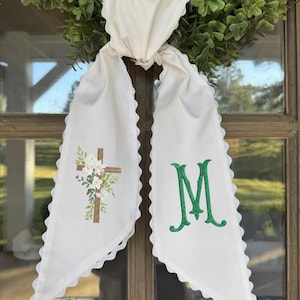 Op de afbeelding: Witte stoffen deurhanger met geschulpte randen, een groene krans en een houten kruis geborduurd met witte bloemen en groen. De rechterkant heeft de groen geborduurde letter "M".