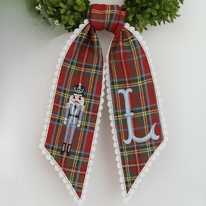 Puede incluir: Bufanda decorativa a cuadros con un ribete de encaje blanco. Un lado presenta un diseño de cascanueces, el otro una letra "L" bordada en azul claro. La bufanda es roja, verde y amarilla.