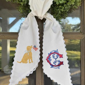 Könnte beinhalten: Weißer Türhänger aus Stoff mit gewelltem Rand, mit einem gestickten Golden Retriever, der eine amerikanische Flagge hält, und einem rot-blauen Monogrammbuchstaben. Der Hänger ist an einem grünen Kranz befestigt.