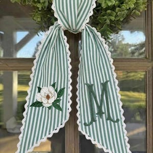 Op de afbeelding: Een groen en wit gestreepte kranssjerp met een witte kanten rand. De sjerp heeft een magnolia bloem borduursel en de letter "M" in groen. De sjerp hangt aan een houten deur.