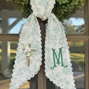 Puede incluir: Un colgador de puerta de tela blanca con un estampado floral verde y bordes festoneados. Un lado presenta una cruz con flores, el otro una "M" verde bordada. El colgador está atado a una corona verde.