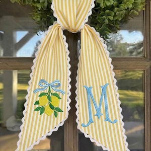 May include: Yellow and white striped fabric door hanger with a scalloped edge. The fabric is adorned with an embroidered lemon and blue bow design, and a blue embroidered letter "M". The door hanger is displayed on a window.