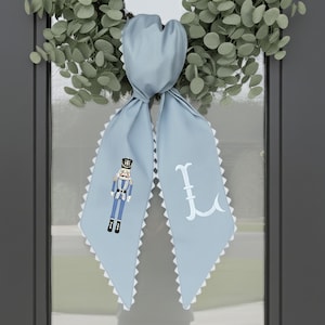 Puede incluir: Un colgador de puerta azul claro con un cascanueces decorativo y la letra "L" bordada. Los bordes están adornados con un ribete festoneado blanco. El colgador de puerta está atado en la parte superior.