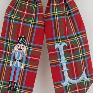 Op de afbeelding: Twee rode geruite kerstsokstrikken met geborduurde details. E&eacute;n strik heeft een blauw-witte notenkrakersoldaat, de andere een lichtblauwe letter "L". De strikken hebben een groen-bruin ruitpatroon.