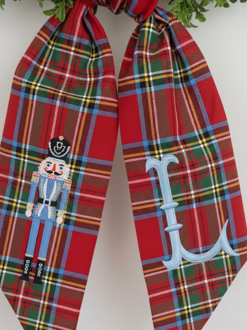 Op de afbeelding: Twee rode geruite kerstsokstrikken met geborduurde details. E&eacute;n strik heeft een blauw-witte notenkrakersoldaat, de andere een lichtblauwe letter "L". De strikken hebben een groen-bruin ruitpatroon.