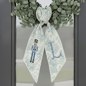 May include: A decorative door bow with a light blue and white toile pattern, featuring a nutcracker and the letter "L". The bow is trimmed with white lace and hangs in front of a green wreath.