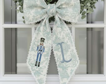 May include: A large decorative bow with a toile pattern in light blue and white, trimmed with white lace. The bow features a nutcracker figure and the letter "L" embroidered on the tails. The bow is hanging on a white door.