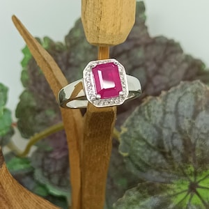 May include: A silver ring featuring a rectangular, vibrant red gemstone, surrounded by a halo of smaller, clear stones. The ring is set against a blurred background of green and purple foliage, highlighting the jewelry's design.