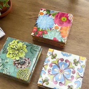 May include: Three square coasters featuring distinct designs: one stack displays a vibrant floral pattern with pink, blue, and orange blooms; another shows green and brown botanical illustrations; the third features a blue and green floral motif.
