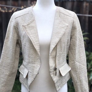 May include: A cream-colored, short-sleeved jacket with a zipper closure and a textured fabric. The jacket has two pockets on the front.