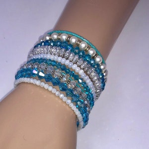 May include: A stack of turquoise and white beaded bracelets with silver accents. The bracelets are layered on a mannequin's wrist.
