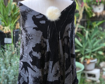 May include: A black and gray patterned velvet shawl with a white fur pom-pom accent. The shawl has a fringe trim.