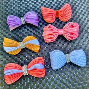 May include: A collection of six colorful fabric bows. The bows are in shades of purple, orange, pink, and blue, with rhinestone embellishments in the center. The bows are arranged on a textured black surface.