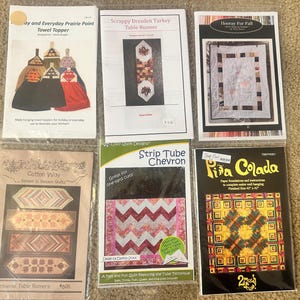May include: A collection of quilting patterns, including designs for towel toppers, table runners, and wall hangings. The patterns feature various styles, from traditional to modern, with colorful illustrations and text descriptions. The finished sizes vary.