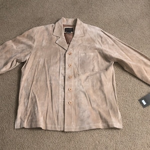 Puede incluir: Una chaqueta de ante marrón claro con cierre de botones y un bolsillo en el lado izquierdo. La chaqueta tiene cuello y mangas largas.