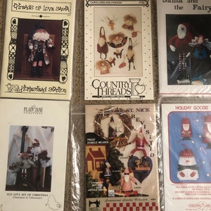 May include: A collection of eight Christmas craft patterns featuring various holiday characters, including Santa, a fairy, a snowman, and a reindeer. The patterns are printed on white paper and are packaged in plastic bags. The patterns are titled "Threads of Love Santa", "Carollers and Friends", "Santa and the Fairy", "Plain Jane Creations", "St. Nick Revisited", "Holiday Goodie Bags", and "#123 Lit'l Bit of Christmas".