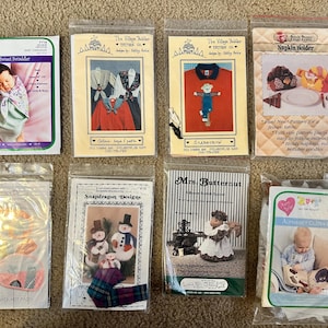 May include: A collection of eight craft patterns for sewing projects. The patterns include a baby swaddler, a scarecrow, a napkin holder, a hot pad, a snowman, a woman in a dress, and a baby's clothing. The patterns are printed on white paper with colorful illustrations.