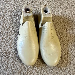 May include: Two off-white shoe lasts, positioned side-by-side, with a rounded toe and a hollowed-out heel. Each last has markings, including size information. The lasts are made of a solid material and show signs of wear.