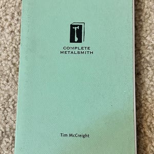May include: A mint green book titled "COMPLETE METALSMITH" with a black hammer and anvil logo. The author's name, Tim McCreight, is printed below the title. The book is lying on a beige carpet.