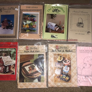 May include: A collection of eight craft patterns for creating various home decor items. The patterns include a gingerbread house, a toilet seat cover, a soft box and basket, Lula's chair, fish sticks, a puff-lid basket, and a doll house. The patterns are printed on white paper with colorful illustrations.