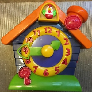 May include: A colorful toy clock shaped like a birdhouse with a rooster on top. The clock face is blue with yellow numbers 1 through 12 and a green hand. The clock has a red button on the side.