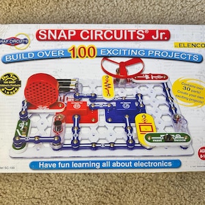 May include: A Snap Circuits Jr. electronics kit with a circuit board, red speaker, and various components. The box reads "Build Over 100 Exciting Projects" and "Have fun learning all about electronics."
