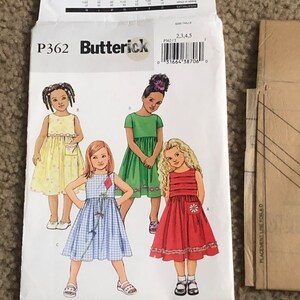 May include: A sewing pattern for girls' dresses. The pattern includes four different dress styles: a yellow dress with white polka dots, a blue and white gingham dress, a green dress with a white collar, and a red dress with a white flower.