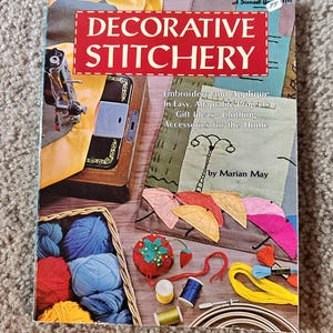 May include: A book titled "Decorative Stitchery" with a red and white title, featuring embroidery and appliqué projects. The image includes a sewing machine, yarn, and embroidery supplies, suggesting a crafting theme.