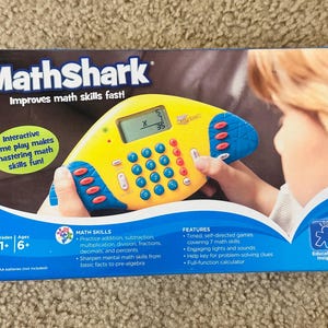 May include: MathShark educational toy. The yellow and blue calculator-like device has buttons and a digital display. The box reads "Improves math skills fast!" and "Interactive game play makes mastering math skills fun!" It is designed for ages 6+.